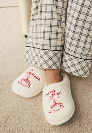 Hvide, bløde hjemmesko med rødt broderet tekst og et motiverende dampende kaffekrus. Kombineret med et tøjstykke med ternet mønster.