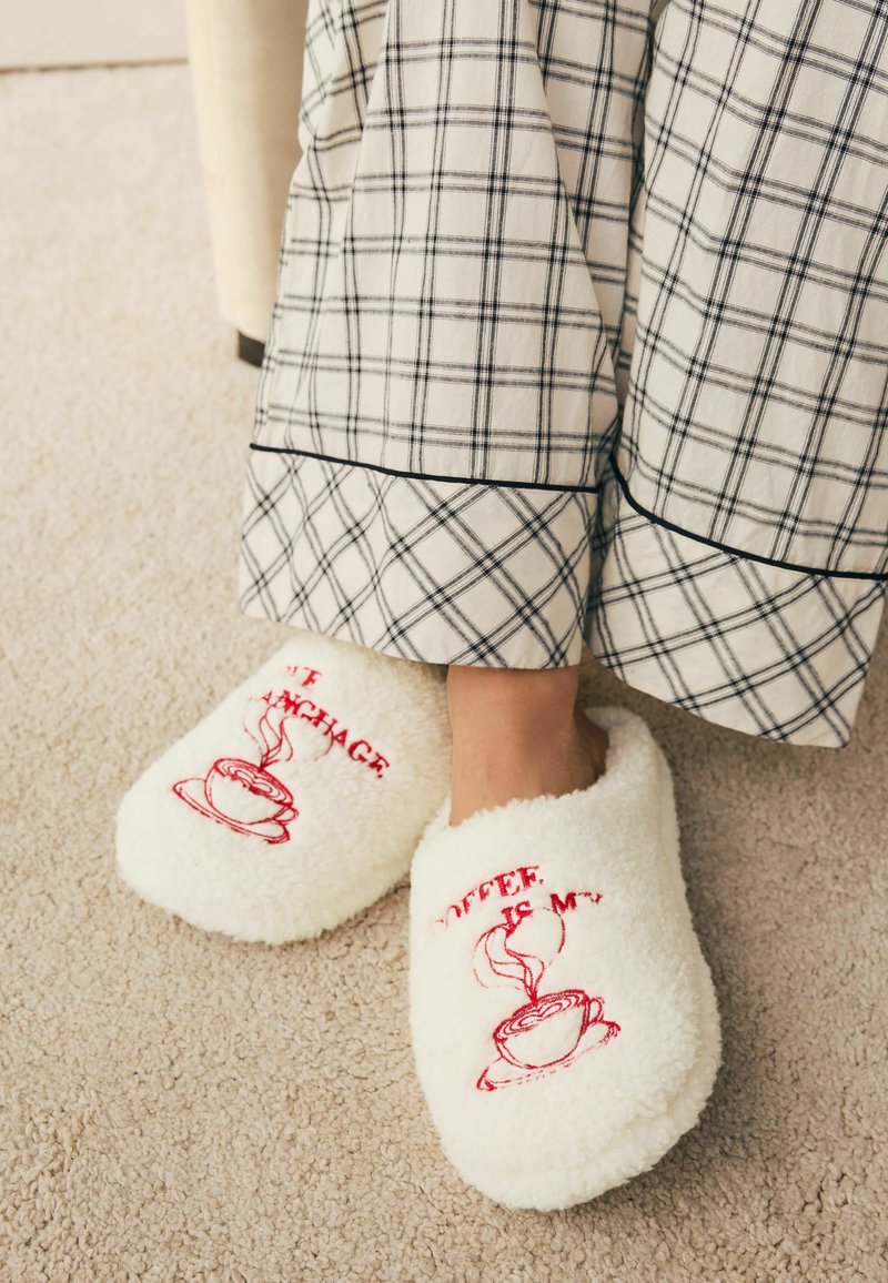 Weiße flauschige Hausschuhe mit roter Stickschrift und einem dampfenden Kaffeetassendesign. Kombiniert mit einem Kleidungsstück im Karierten Muster.