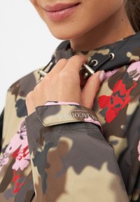 Kurtka w wzór kamuflażu w odcieniach brązu z różowymi i czerwonymi kwiatowymi akcentami. Posiada kaptur ze sznurkiem i metkę z logo na mankiecie.