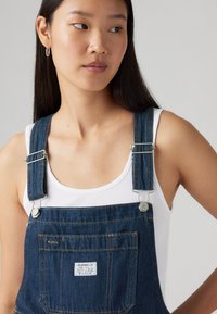 Dongeribukser i mørkeblå, med justerbare stropper, sølvfarvede detaljer og en frontlomme med et mærke. Båret over en hvid tanktop.