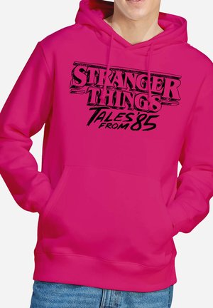 Person trägt einen leuchtend pinken Hoodie mit schwarzem Schriftzug "Stranger Things Tales from 85" auf der Vorderseite und die Hände in der Kängurutasche.
