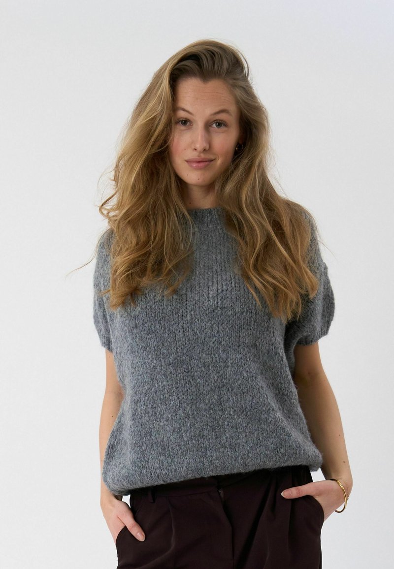 Jeune femme aux longs cheveux blonds portant un pull court gris lâche en tricot à manches courtes et un pantalon foncé, mains dans les poches, fond neutre.