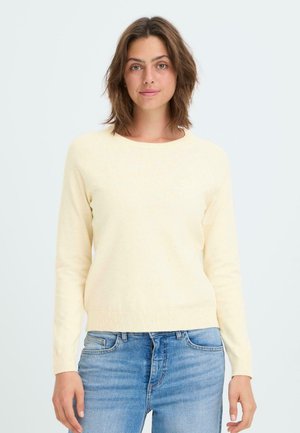 Maglione lavorato a maglia di un giallo chiaro con maniche lunghe e collo rotondo, abbinato a jeans di denim blu medio, su uno sfondo semplice.