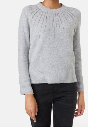 Pull gris clair à manches longues avec motif rayons de soleil en perles sur la poitrine, associé à un jean noir, porté par une personne sur un fond uni.