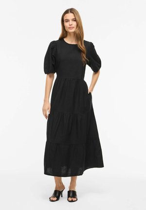 Kvinde står iført en sort lagdelt midi-kjole med puffede korte ærmer og sorte åben tå hælede mule, mod en ensfarvet hvid baggrund.