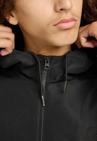 Persona che alza il colletto di un hoodie nero con zip, con un logo ricamato vicino all'angolo in basso a destra.