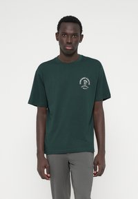 Mees seisab ettepoole, seljas tumesinine T-särk, millel on valge kohvipoti logo ja tekst vasakul rinnal, koos hallide pükstega.