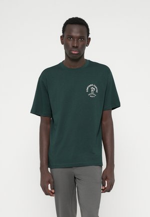 Ein Mann steht mit Blick nach vorne da, trägt ein dunkelgrünes T-Shirt mit einem weißen Kaffeekannen-Logo und Text auf der linken Brust, kombiniert mit grauen Hosen.