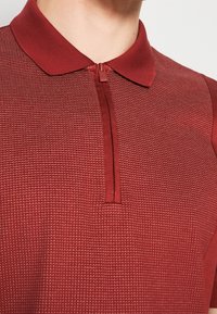 Gros plan d'une personne portant un polo rouge texturé avec un col zippé devant un fond uni.