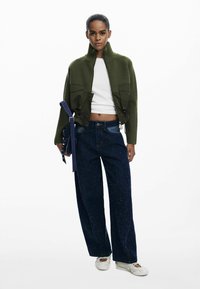 Giacca accorciata verde oliva con trama a costine e due tasche frontali, abbinata a pantaloni navy a gamba larga con una leggera lucentezza. Scarpe bianche.
