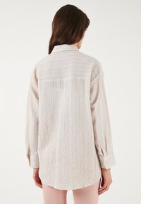 Light beige textured shirt with a button-down collar, long sleeves, and a relaxed fit, featuring subtle horizontal pleats and side slits.