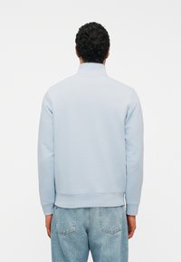 Homme aux cheveux bouclés portant un sweat-shirt bleu clair et un jean bleu, vu de dos contre un fond blanc uni.