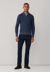 Maglione blu navy a maglia con colletto a mezza zip, indossato sopra una camicia azzurro chiaro. Abbinato a jeans denim scuri e scarpe in suede marroni.