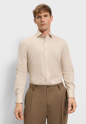 Man draagt een beige gestreept overhemd met lange mouwen, ingestopt in een hooggetailleerde bruine broek, tegen een effen achtergrond.