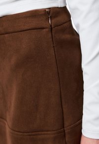 Gros plan sur une jupe en daim marron avec une fermeture éclair latérale portée avec un haut blanc à manches longues.