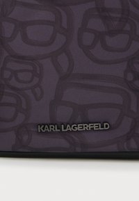 Tamsus audinys su pakartotiniais dideliais akinių kontūrais ir mažu juodu metaliniu "KARL LAGERFELD" logotipu apatiniame centre.