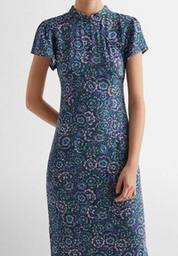 Kukkakuosisella mekolla, jossa on lyhyet hihat. Mekossa on tummalla taustalla sinisen ja vihreän sävyjä, ja se on valmistettu sileästä kankaasta.