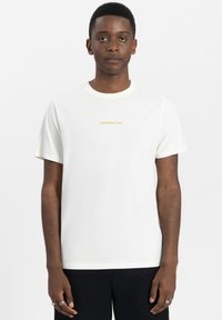 Witte katoenen t-shirt met een ronde hals en korte mouwen. De voorkant toont "TOMORROWLAND" in kleine, gouden letters.