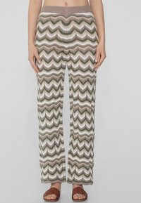 Gehaakte broek met wijde pijpen in een zigzagpatroon in groen, wit en beige. Kenmerkt zich door een bruine elastische tailleband en een open haakstructuur.