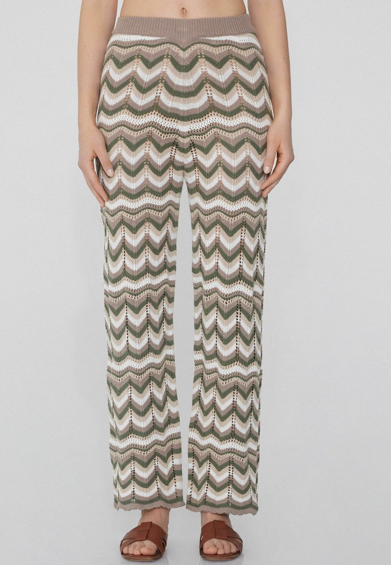 Gehaakte broek met wijde pijpen in een zigzagpatroon in groen, wit en beige. Kenmerkt zich door een bruine elastische tailleband en een open haakstructuur.