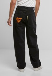 Schwarze Denim-Hosen mit lockerer Passform, mit gelb-rot gesticktem "Home Boy"-Text auf der Gesäßtasche und einem Reißverschlusstaschen-Detail.