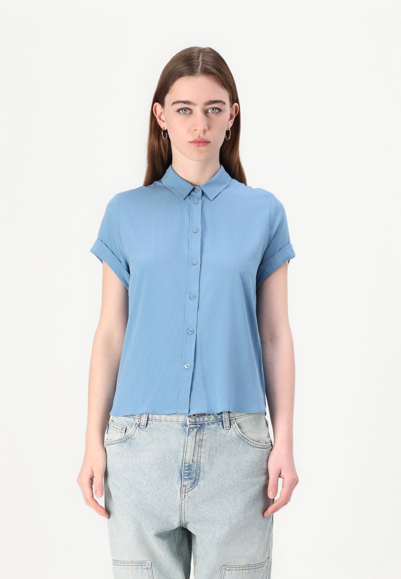 Light blue short-sleeve button-up shirt made from a smooth fabric with a collared neck and rolled sleeves, paired with loose denim pants.