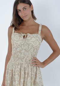 Beżowa sukienka w wzory z marszczoną górą, cienkimi ramiączkami i szczegółem w postaci wiązania z przodu. Posiada zwiewny dół i subtelny kwiatowy wzór.