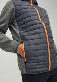 Puchowa kurtka z granatowym pikowanym przodem i szarymi teksturowanymi rękawami. Posiada pomarańczowy zamek i akcenty, które dodają kontrastu i detalu.