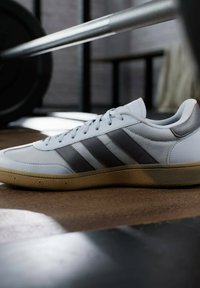 Witte sneaker met grijze strepen, gestructureerde synthetische bovenkant, rubberen gum zool, ronde neus en platte veters, gepositioneerd in een gymomgeving.
