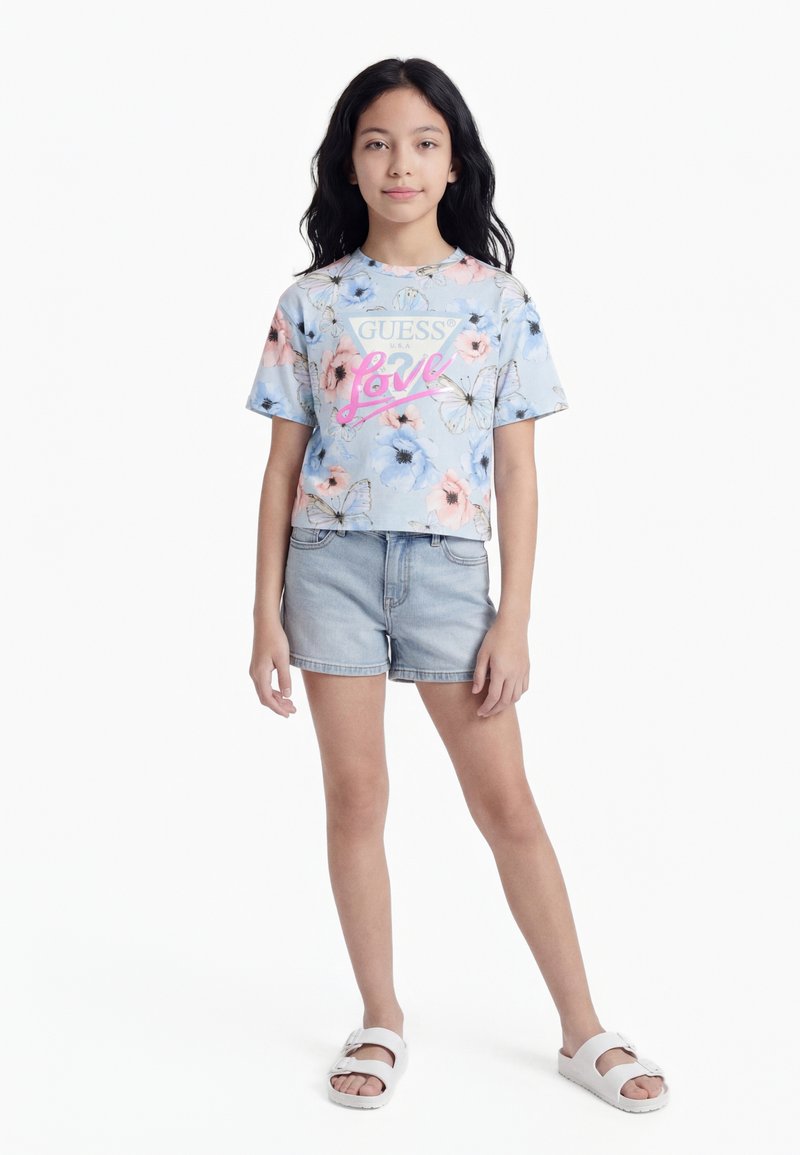 Κορίτσι που στέκεται φορώντας μια μπλούζα Guess με λουλουδάτο σχέδιο, τζιν σορτς και λευκά σανδάλια, πάνω σε απλό λευκό φόντο.