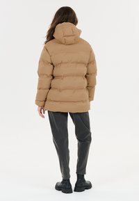 Person med langt hår iført en beige pufferjakke, mørke tapered bukser og sorte chunky støvler, der står på en hvid baggrund med ryggen til.