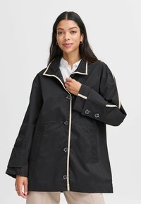 Zwarte regenjas met beige biezen, voorzien van een kraag, knoopsluiting en twee voorzakken. Gemaakt van duurzaam, glad materiaal.