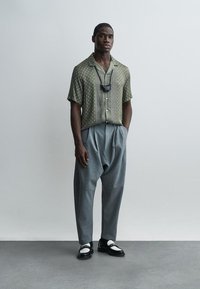 Mann iført mønstret kortermet skjorte, løse grå plisserte bukser, svart-hvite loafers og svart ansiktsmaske hengende fra nakken, stående mot en ensfarget vegg.