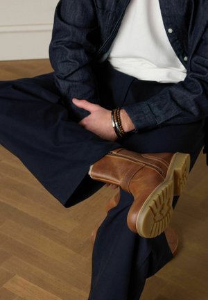 Persoon zittend met gekruiste benen, draagt bruine leren laarzen, donkerblauwe broek, een wit shirt en een donkerblauwe spijkerjasje met armbanden.