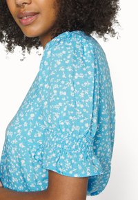 Blusa turquesa com padrão floral, mangas bufantes e punhos elásticos, apresentando uma textura de tecido suave e leve.
