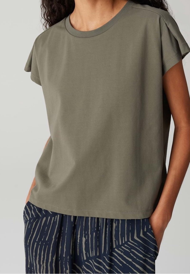 Femme portant un haut à manches courtes vert olive et un pantalon bleu marine avec des rayures claires verticales et diagonales, mains dans les poches.