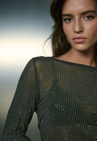 Top verde scuro trasparente a maniche lunghe decorato con borchie metalliche su tutta la superficie, caratterizzato da una scollatura ampia e una texture liscia.