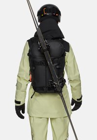 Schwarzer Rucksack mit mehreren Fächern, verstellbaren Trägern und angehängter Skiausrüstung, getragen über einer hellgrünen Außenschicht.