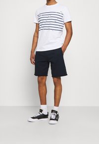 Weißes T-Shirt mit navyblauen horizontalen Streifen und Palmbaum-Stickerei, kombiniert mit navyblauen Shorts und schwarzen Sneakern.