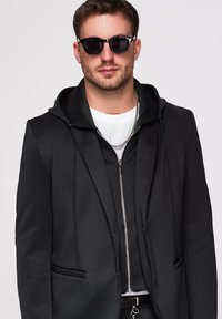Homme portant des lunettes de soleil noires, un sweat à capuche noir zippé sous un blazer noir, une chemise blanche et une chaîne argentée.