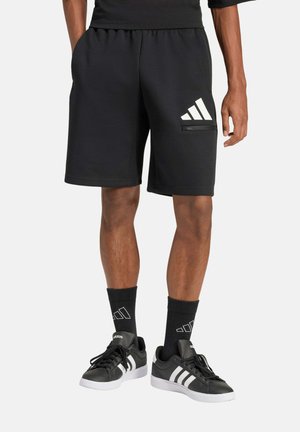 Hombre con pantalones cortos deportivos negros con logo blanco, calcetines negros y zapatillas negras con rayas blancas, de pie sobre fondo liso.