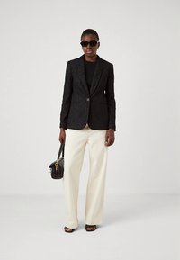 Blazer negro texturizado con un solo botón, combinado con una blusa negra, pantalones anchos color crema y un bolso negro. La modelo lleva gafas de sol.