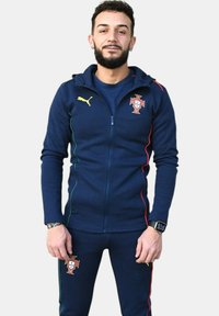 Chándal navy hecho de tela suave, con chaqueta con cremallera que presenta el logo amarillo de Puma y el emblema de Portugal, acentuado con rayas rojas y verdes.