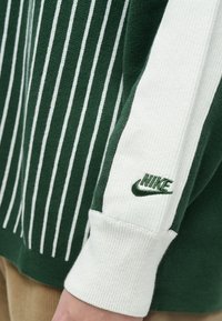 Zöld és fehér Nike pulóver függőleges csíkokkal, bordázott mandzsettákkal és zölden hímzett logóval az ujján. Puha anyag textúra.