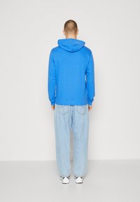 Sudadera azul con bolsillo canguro y capucha con cordón, combinada con vaqueros holgados de un azul claro y zapatillas blancas, vista desde atrás.