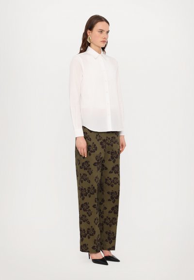 Chemise blanche à boutons et manches longues associée à un pantalon large vert avec un motif floral sombre. Des escarpins noirs à bout pointu complètent le look.