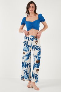 Sieviete stāv, valkājot zilu rušētu īso topu ar pūkainām piedurknēm un augsta vidukļa baltas bikses ar zilu un brūnu lapu rakstu, bēšas kurpes ar papēdi.