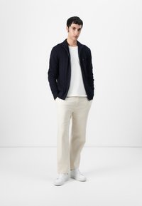 Marineblå kabelstrikket zip-sweater over en hvit t-skjorte, kombinert med kremfargede vide bukser og hvite sneakers. Enkle, rene linjer og teksturer.