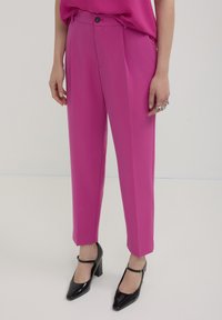 Pantaloni fucsia con vestibilità sartoriale, dotati di chiusura a bottoni e pieghe, abbinati a scarpe nere con tacco e una blusa coordinata.