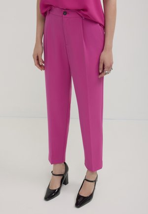 Pantalons fuchsia avec une coupe ajustée, dotés d'une fermeture à boutons et de plis, associés à des chaussures à talons noires et un haut assorti.
